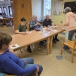 Več oseb sedi za mizo in ima pred seboj pijačo ter pecivo na prtičkih.