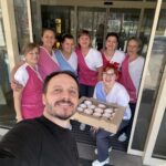 Skupina zaposlenih stoji pred vhodom v dom in se fotografira; ena sodelavka drži škatlo s krofi, moški v ospredju pa dela skupinski selfi.