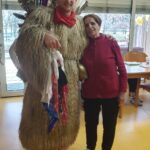 Kurent stoji ob stanovalki v rdečem puloverju; objeta pozirata v skupnem prostoru doma.