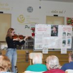 Oseba stoji in igra violino pred sedečo skupino oseb v prostoru z razstavnim panojem.