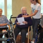 Sedeča oseba bere z lista v mikrofon, ob njej stoji druga oseba, okoli pa sedijo druge osebe.