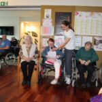 Več oseb sedi na invalidskih vozičkih ali stolih, ena oseba bere z lista v mikrofon.