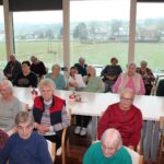 Skupina starejših oseb sedi za mizami in posluša osebo, ki govori v mikrofon.