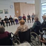 Oseba stoji v krogu sedečih oseb in bere z lista v mikrofon.