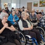 Skupina starejših oseb sedi na invalidskih vozičkih ali stolih in posluša dogajanje v prostoru.