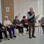 Moški stoji, drži mikrofon in bere z lista pred sedečo skupino oseb.