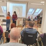 Dve dekleti stojita pred projekcijskim platnom in bereta besedilo občinstvu, ki sedi na stolih in invalidskih vozičkih.