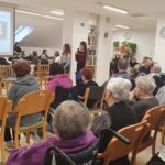 Dekle z mikrofonom govori pred zbranim občinstvom, na platnu je prikazana predstavitev.