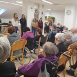 Skupina deklet stoji pred občinstvom, ena govori v mikrofon, ostale držijo liste z besedilom.