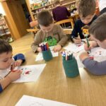 Več otrok sedi za mizo in barva risbe z barvnimi flomastri, na mizi so lončki s pisali.