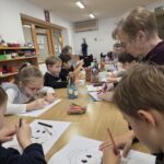 Otroci in starejše osebe sedijo okoli mize in skupaj rišejo ter barvajo, na mizi so barvice, papir in kozarci.