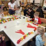 Otroci in starejše osebe sedijo za mizo in izdelujejo papirnate verige iz barvnega papirja, na mizi so škarje in lepilo.