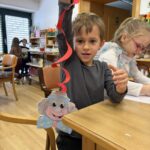 Otrok stoji ob mizi in drži papirnato pustno dekoracijo z obrazom, nanjo je pritrjen rdeč papirni trak.