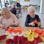 Dve osebi sedita za mizo in oblikujeta barvne papirnate elemente, na mizi so zloženi kosi rdečega, rumenega in belega papirja.