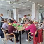 Več oseb sedi okoli miz v večnamenskem prostoru in skupaj ustvarja z barvnim papirjem, ena oseba stoji in razdeljuje material.