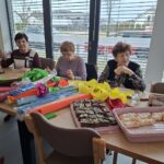 Tri osebe sedijo za mizo ob oknu, pred njimi so barvni papir, škarje in pladnji s pecivom ter kozarci z vodo.