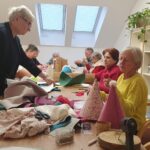 Več oseb sedi in stoji okoli mize ter iz blaga in papirja izdeluje okrasne predmete.