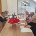 Skupina žensk sedi za mizo in riše ter izrezuje oblike iz papirja.