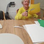 Ženska sedi za mizo in s škarjami izrezuje obliko iz rumenega papirja.