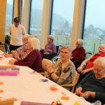 Skupina oseb sedi za mizo in iz barvnega papirja izdeluje okraske v prostoru z velikimi okni.