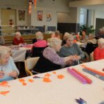 Več oseb sedi za mizami in izrezuje barvni papir v jedilnici z razdelilnim pultom v ozadju.