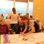 Oseba pomaga sedeči osebi pri oblikovanju papirnatega okraska za mizo z ustvarjalnim materialom.