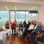 Oseba pripravlja jed v posodi na mizi, medtem ko druge osebe sedijo in opazujejo v skupnem prostoru.