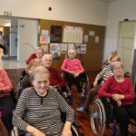 Več oseb v invalidskih vozičkih sedi v polkrogu v skupnem prostoru, nekatere v rokah držijo papirnate trakove.