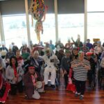 Skupina oseb v različnih pustnih kostumih pozira skupaj v notranjem prostoru pred velikimi okni.