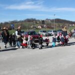 Skupina otrok in nekaj odraslih hodi po parkirišču na prostem v bližini parkiranih vozil in travnatih površin.