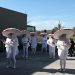 Več oseb v enakih belih pustnih kostumih z velikimi okroglimi pokrivali stoji na prostem pred stavbo.