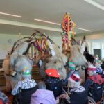 Več oseb v kosmatih pustnih kostumih nastopa pred sedečimi osebami v notranjem prostoru z visečimi barvnimi okraski.