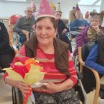 Stanovalka sedi na vozičku, nosi rožnato pustno kapo in v rokah drži veliko papirnato rožo, v ozadju pa sedijo tudi drugi stanovalci z različnimi pustnimi klobuki.