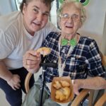 Zaposlena in stanovalka z zelenimi pustnimi dodatki pozirata s krofi v roki.