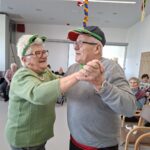 Stanovalca z zelenimi dodatki plešeta v skupnem prostoru, medtem ko drugi sedijo ob strani.