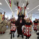 Kurenti in spremljevalke v rdeče-črnih kostumih hodijo skozi prostor, okrašen s pisanimi trakovi.