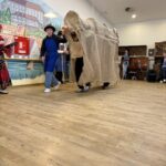 Skupina oseb v kostumih hodi po prostoru, ena oseba vodi veliko pokrito lutko ali rekvizit.