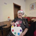 Ženska sedi za mizo, nosi pokrivalo in v rokah drži papirnat rekvizit v obliki palete z barvnimi krogi.