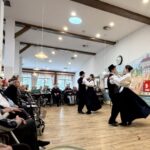 Pari oseb v tradicionalnih oblačilih plešejo v prostoru, ob strani jih spremlja sedeče občinstvo.