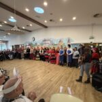 Skupina oseb v kostumih stoji v vrsti v prostoru, ob strani jih opazuje in jim ploska občinstvo.