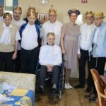 Skupina stanovalcev z zlatimi kronami pozira v skupnem prostoru; ena oseba sedi na invalidskem vozičku.