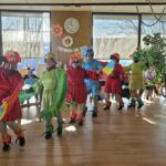 Skupina oseb v pisanih kostumih ptic pleše v krogu, medtem ko stanovalci sedijo ob strani in opazujejo.