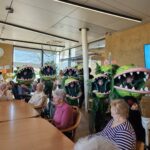 Osebe v zelenih kostumih z velikimi odprtimi usti vstopajo v jedilnico med sedeče stanovalce.