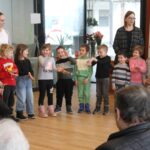 Skupina otrok stoji v krogu in izvaja skupinsko dejavnost, ob njih stojita dve odrasli osebi.