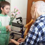 Otrok podaja cvetje sedeči osebi ob hojci.