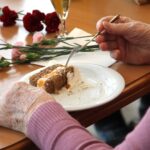 Roka osebe drži vilico nad kosom torte na krožniku, na mizi so rože in kozarec pijače.