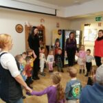 Otroci in odrasle osebe stojijo v krogu v notranjem prostoru in se držijo za roke.