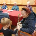 Otrok stoji pred sedečo osebo in ji izroča cvet, v ozadju so mize s pogrinjkom in druge osebe.