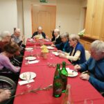 Več oseb sedi za dolgo mizo s pogrinjkom in praznuje ob obroku, na mizi so krožniki, kozarci in cvetje.