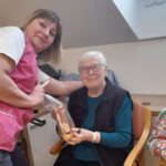 Ženska stoji ob sedeči starejši ženski in ji izroča zavit paket.